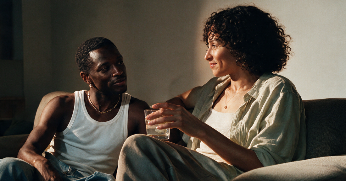 Two people sitting together on a sofa in warm evening light, one holding a glass of clear sparkling water